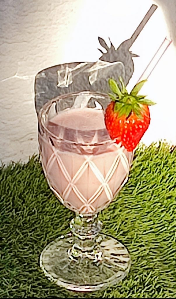 Copa transparente con batido rosa sobre césped con fondo de pared blanca y sombra marcada por la luz natural.