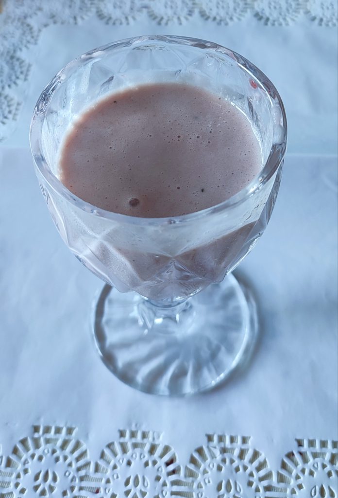 Copa de cristal con batido rosa sobre mesa interior con blonda blanca, mostrando burbujas y textura cremosa.