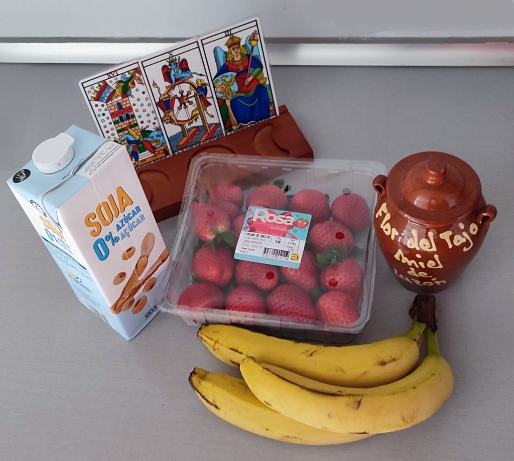 Plátanos, fresas, miel y bebida vegetal junto a expositor de tarot con las cartas La Torre, La Rueda y La Emperatriz sobre encimera gris.